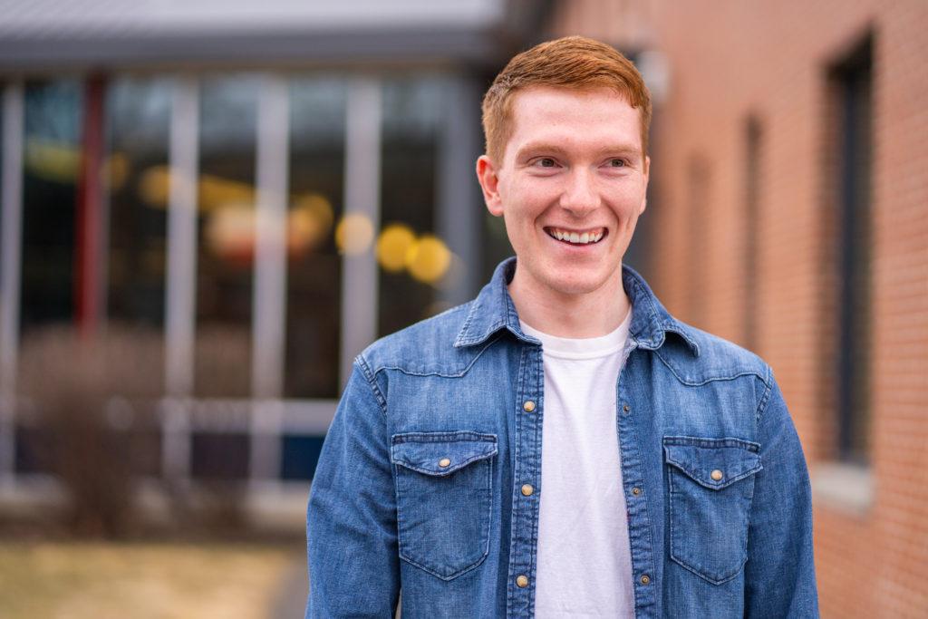 person with blue jean shirt and red hair smiles to side of the camera with building with big tall windows in the background